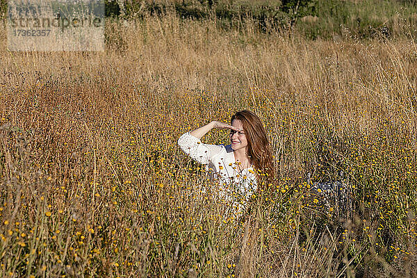 Lächelnde junge Frau  die ihre Augen abschirmt  während sie auf einem Feld wegschaut