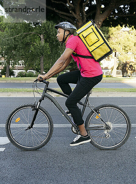 Männlicher Zusteller mit gelbem Rucksack fährt mit dem Fahrrad auf der Straße