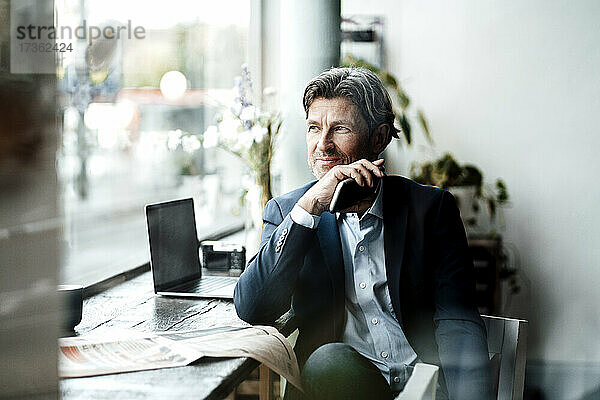 Lächelnder Geschäftsmann  der sein Smartphone hält und durch das Fenster schaut  während er in einem Café am Tisch sitzt