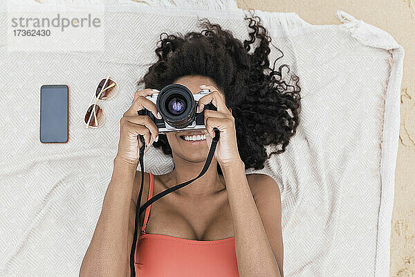 Lächelnde junge Frau  die durch die Kamera fotografiert  während sie auf einem Handtuch am Strand liegt