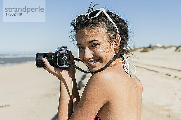 Lächelnde junge Frau mit Kamera am Strand stehend an einem sonnigen Tag