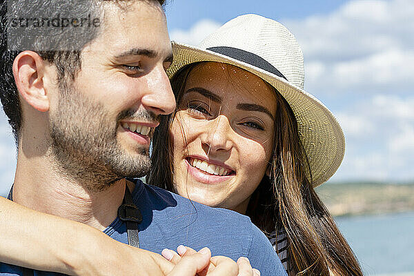 Lächelnde Frau mit Hut umarmt ihren Freund an einem sonnigen Tag