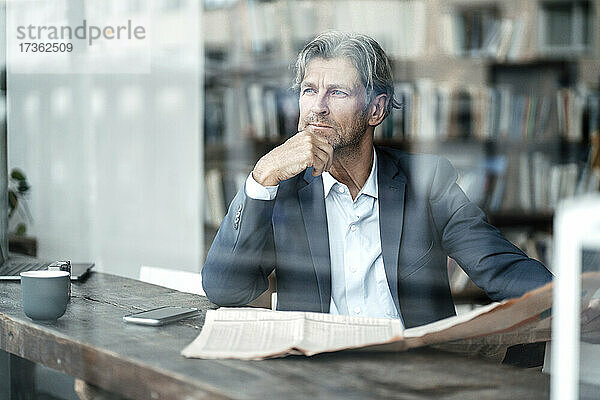 Nachdenklicher männlicher Berufstätiger  der mit der Hand am Kinn in einem Café sitzt  gesehen durch ein Glasfenster