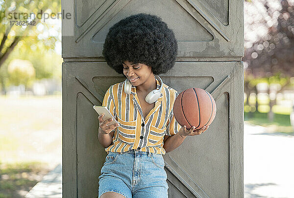 Lächelnde Frau  die einen Basketball hält und ein Mobiltelefon benutzt