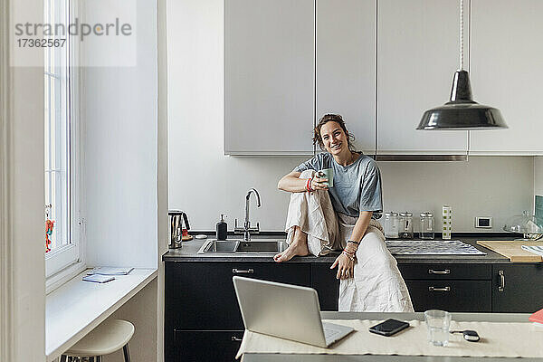 Lächelnde Freiberuflerin  die eine Kaffeetasse hält  während sie auf dem Küchentisch sitzt