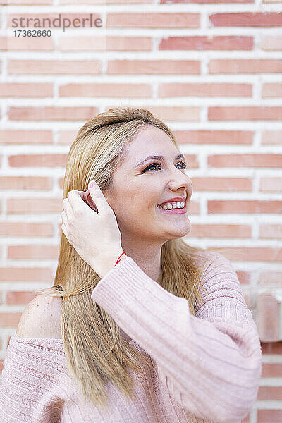 Junge Frau mit Hand im Haar lächelnd vor Backsteinmauer