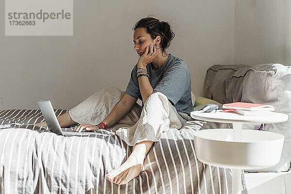 Weibliche Fachkraft  die einen Laptop benutzt  während sie mit der Hand am Kinn im Schlafzimmer sitzt