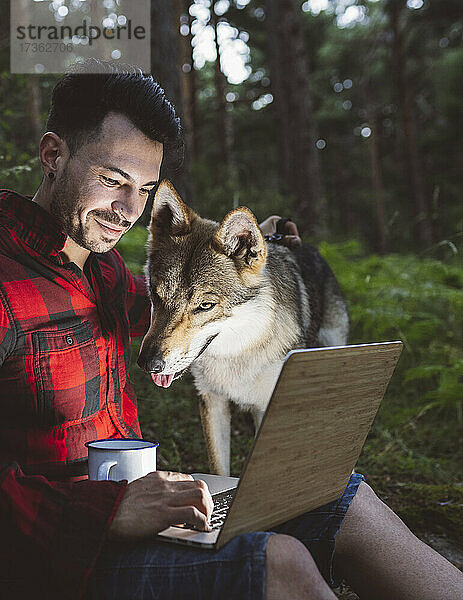 Männlicher Wanderer mit Kaffeetasse und Laptop  während er neben einem Hund im Wald sitzt