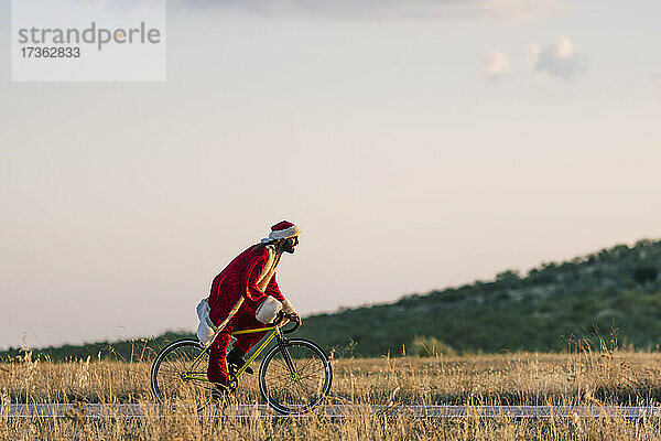 Junger Mann im Weihnachtsmannkostüm fährt bei Sonnenuntergang Fahrrad auf der Straße