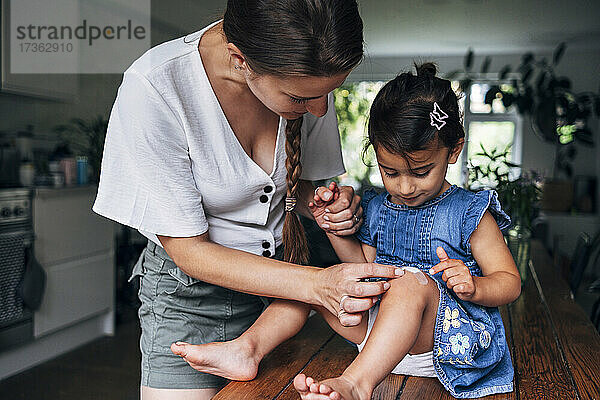 Mutter hält Hand  während sie zu Hause einen Verband auf das Knie ihrer Tochter legt