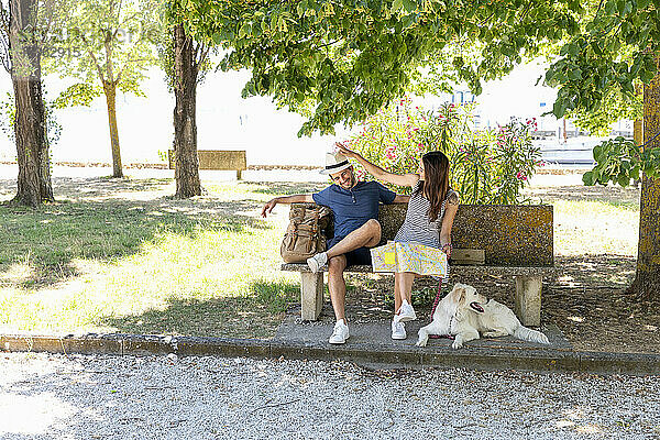 Touristenpaar mit Landkarte und Rucksack sitzt mit Hund auf einer Bank