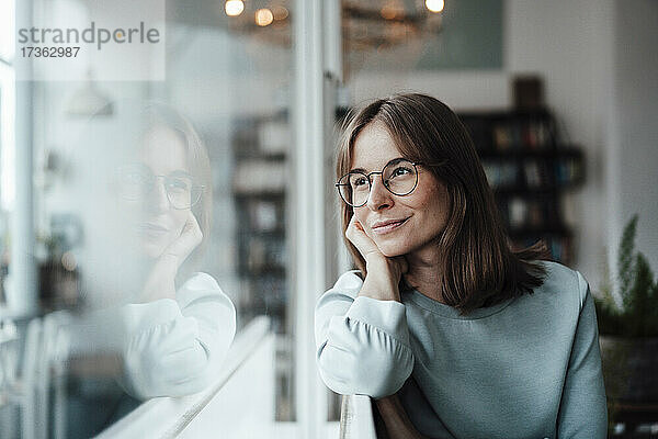 Lächelnde nachdenkliche Frau mit braunen Haaren  die durch das Fenster schaut  während sie in einem Café sitzt