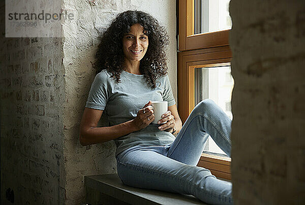 Lächelnde weibliche Fachkraft  die eine Kaffeetasse hält  während sie auf der Fensterbank sitzt