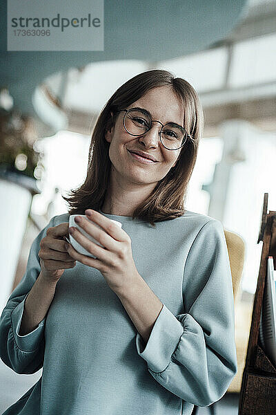 Lächelnde junge Frau mit braunen Haaren hält Kaffeetasse in Café