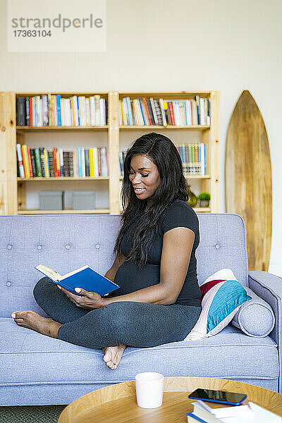 Lächelnde junge schwangere Frau  die auf dem Sofa im Wohnzimmer sitzt und ein Buch liest