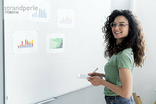 Lächelnde Geschäftsfrau  die ein digitales Tablet hält  während sie an einem Whiteboard im Büro steht