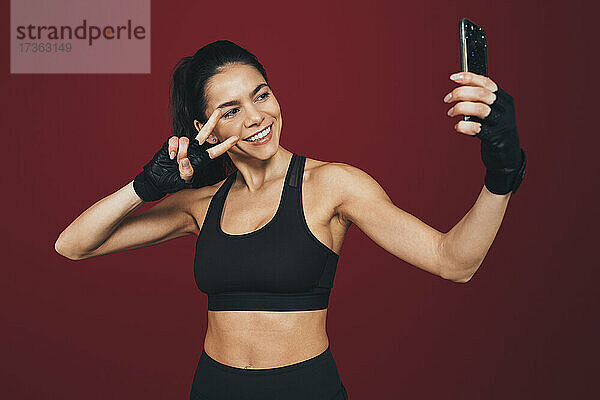 Lächelnde Sportlerin gestikuliert Friedenszeichen bei der Aufnahme von Selfie durch Handy
