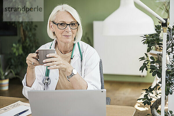 Ältere Ärztin hält Kaffeetasse am Schreibtisch
