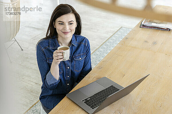 Berufstätige Frau trinkt Kaffee und sitzt am Laptop im Büro