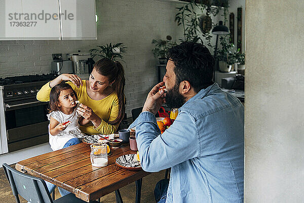 Frau füttert ihre Tochter mit Essen  während sie mit einem Mann am Tisch sitzt