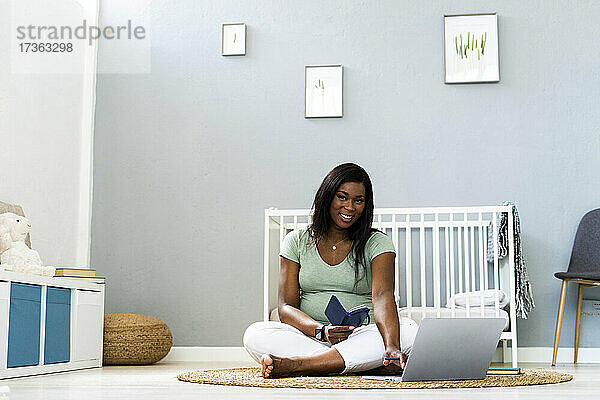 Lächelnde junge schwangere Frau im Schneidersitz mit Laptop im Schlafzimmer