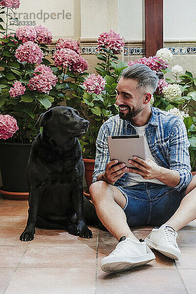 Lächelnder Geschäftsmann mit digitalem Tablet  der seinen Hund ansieht  während er im Hinterhof auf dem Boden sitzt