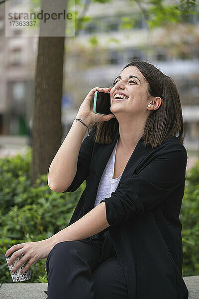 Glückliche junge Frau  die im Park sitzt und mit ihrem Handy telefoniert