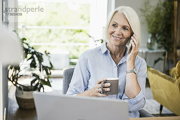 Lächelnde Geschäftsfrau  die eine Tasse hält  während sie im Büro zu Hause mit einem Smartphone spricht