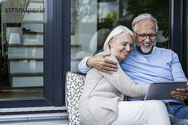 Lächelnder Mann  der ein digitales Tablet benutzt  während er mit dem Arm um eine Frau auf dem Balkon sitzt