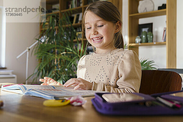 Lächelndes Mädchen  das beim Lernen zu Hause ein Buch auf dem Tisch betrachtet