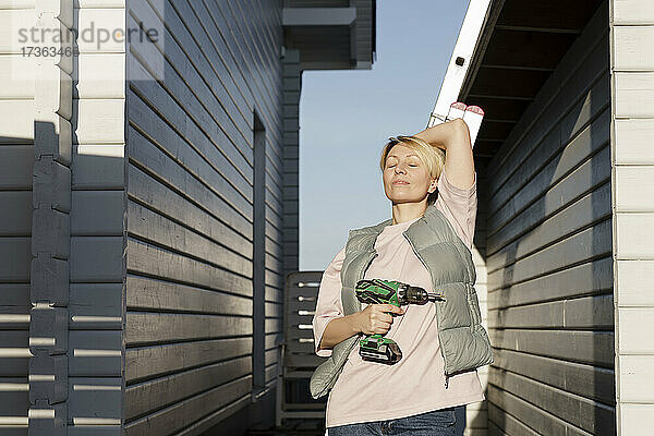 Frau mit geschlossenen Augen lehnt sich an eine Leiter und hält eine Bohrmaschine in der Nähe eines Hauses