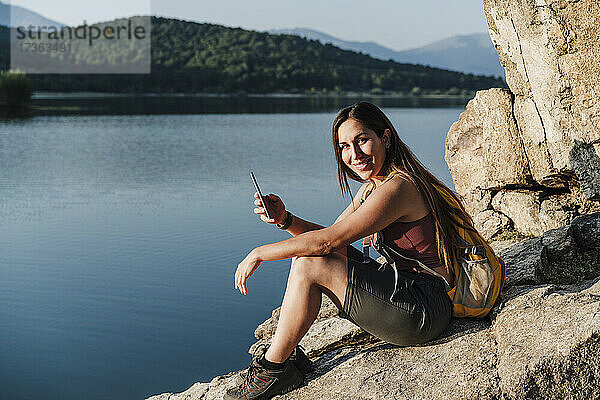 Lächelnde Wanderin mit Handy in der Hand auf einem Felsen am See sitzend