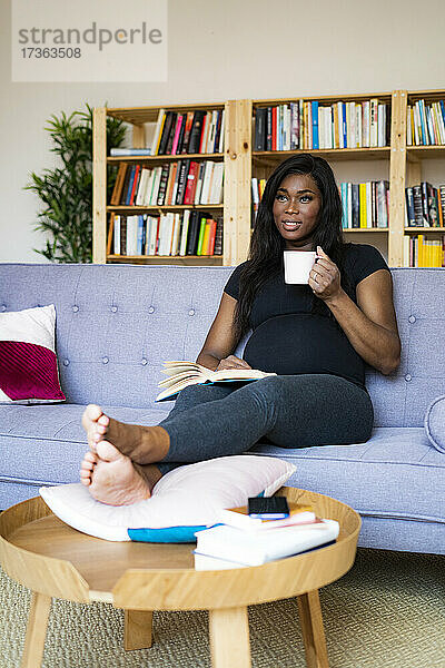 Nachdenkliche schwangere Frau hält eine Kaffeetasse  während sie zu Hause auf dem Sofa sitzt