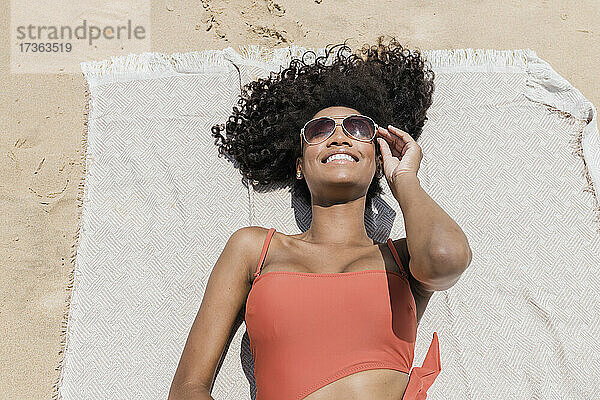 Lächelnde junge Frau mit lockigem Haar und Sonnenbrille  die auf einem Strandtuch liegt