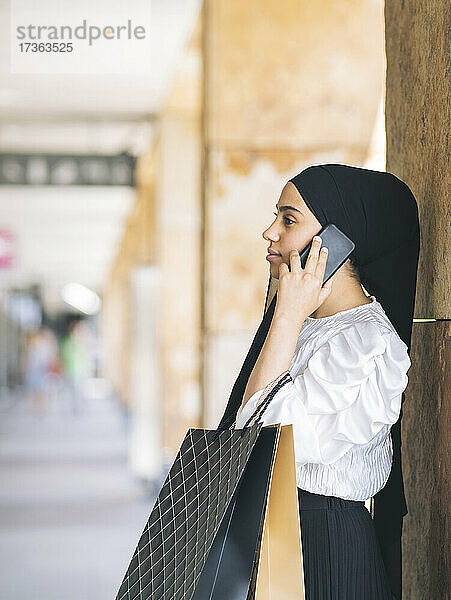 Frau mit Einkaufstüten  die mit einem Smartphone an einer Säule telefoniert