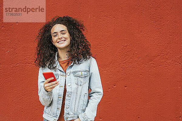 Lächelnde Frau hält ihr Smartphone vor einer roten Wand