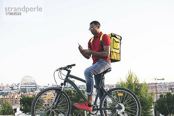 Mann mit Sonnenbrille  der im Stehen mit dem Fahrrad eine Textnachricht über sein Smartphone verschickt