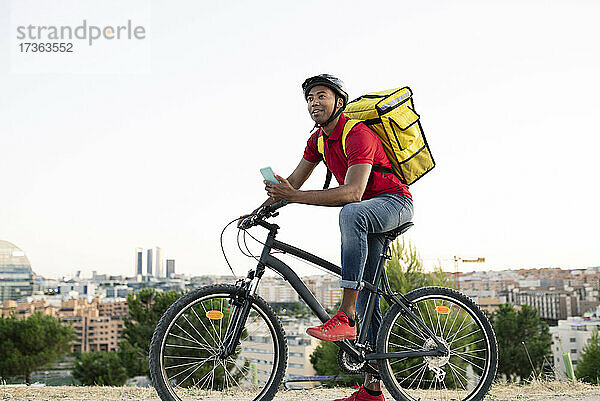 Männlicher Zusteller  der ein Smartphone hält und wegschaut  während er sich auf ein Fahrrad stützt