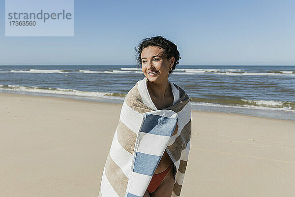 Lächelnde Frau  eingewickelt in ein Handtuch  schaut weg  während sie am Strand steht