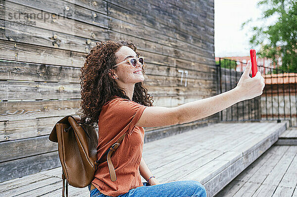 Lächelnde junge Frau mit Rucksack nimmt Selfie durch Smartphone