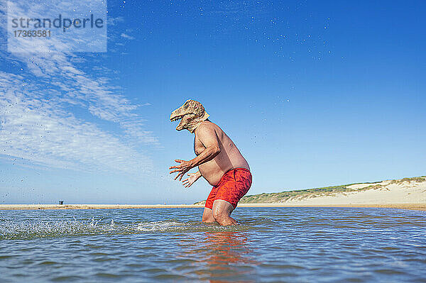 Mann mit Eidechsenmaske gestikuliert im Meer am Strand