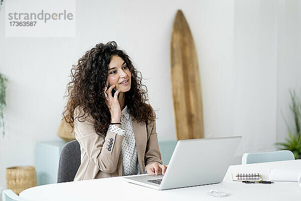 Geschäftsfrau mit Laptop am Schreibtisch  die mit einem Mobiltelefon im Büro spricht
