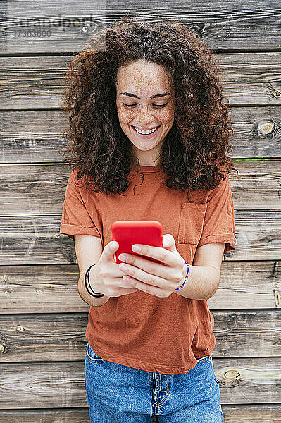 Lächelnde Frau mit lockigem Haar  die ein Smartphone vor einer Holzwand benutzt