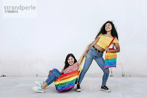 Lächelnde lesbische Frau  die ihre in einer Regenbogentasche sitzende Freundin auf dem Fußweg vor einer weißen Wand zieht