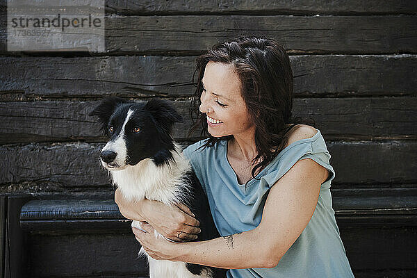 Glückliche Frau  die ihren Border Collie umarmt  während sie vor einer Hütte sitzt