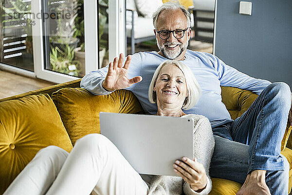 Lächelnder Mann winkt mit der Hand während eines Videoanrufs über einen Laptop  während er mit einer Frau auf dem Sofa sitzt