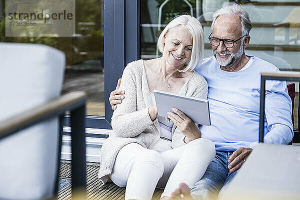 Lächelnde Frau  die ein digitales Tablet benutzt und neben einem Mann auf dem Balkon sitzt