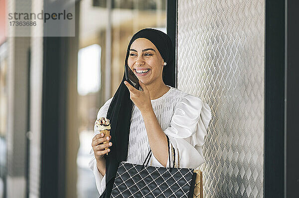 Glückliche Frau mit Eiscreme  die vor einem Fenster mit ihrem Smartphone telefoniert