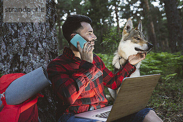 Mann mit Laptop  der mit seinem Handy telefoniert  während er neben einem Hund im Wald sitzt