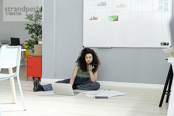 Geschäftsfrau  die mit einem Mobiltelefon spricht  während sie im Büro an einem Laptop arbeitet